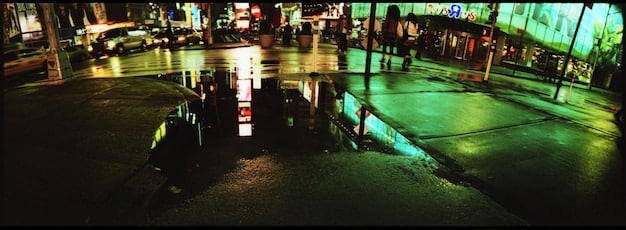 A wide, panoramic panel from 'From' showcasing a detailed cityscape at night, with neon lights reflecting in the rain-slicked streets, creating a sense of urban isolation and mystery.
